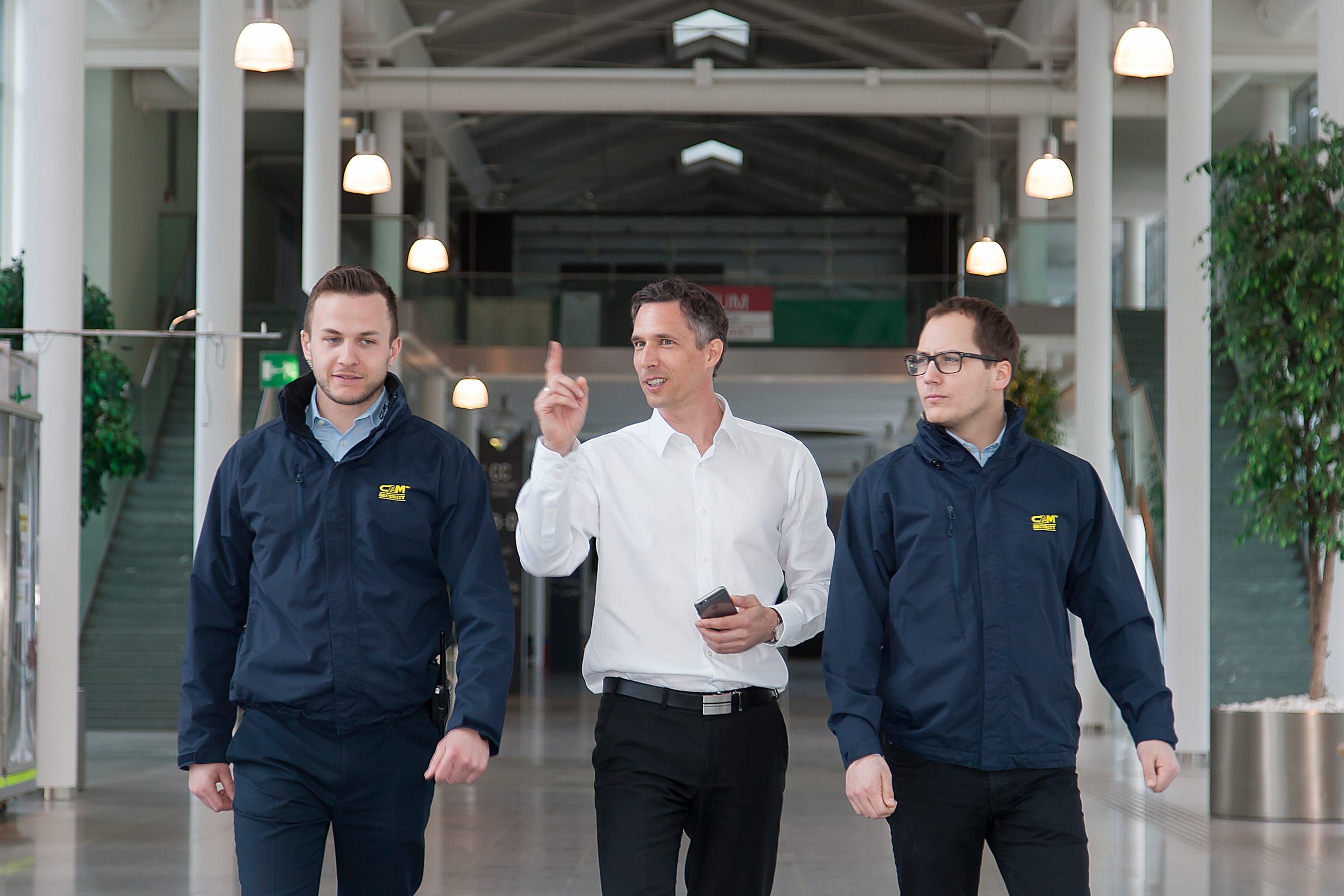 Drei Teammitglieder von CAM Security gehen durch ein Foyer, zwei davon in dunkelblauen CAM-Jacken